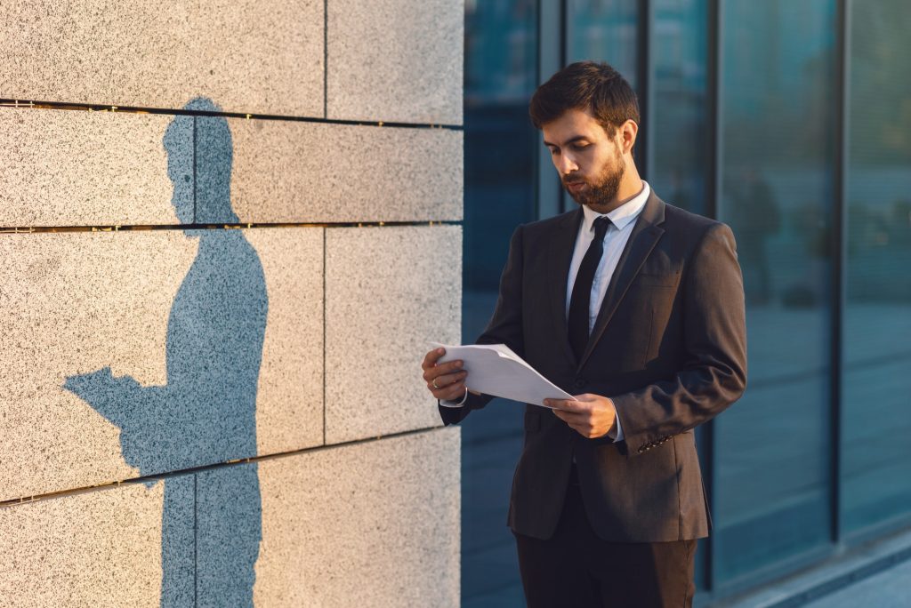 Serious young businessman in a classic suit reading advantageous business document in paper